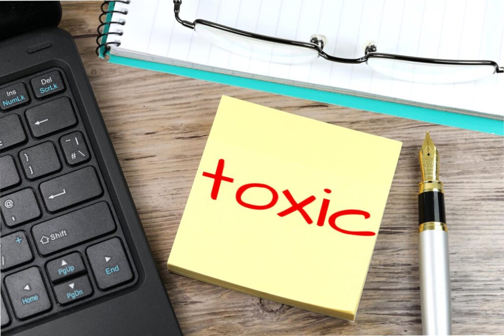 Desktop with keyboard, pen, notebook, classes and a yellow post-it note in the center. The word "toxic" is written in red on the post-it.