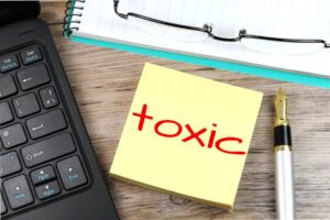 Desktop with keyboard, pen, notebook, classes and a yellow post-it note in the center. The word "toxic" is written in red on the post-it.