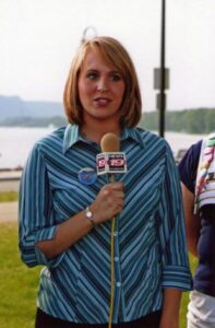 Throwback picture of Sara stands in the grass near a river holding a TV microphone with the insignia for WXOW channel 19 for a live shot. She's wearing blue striped button down collar shirt and is in the middle of addressing a camera which isn't visible in the picture.