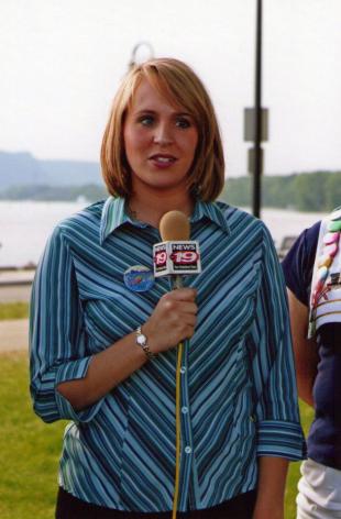 Throwback picture of Sara stands in the grass near a river holding a TV microphone with the insignia for WXOW channel 19 for a live shot. She's wearing blue striped button down collar shirt and is in the middle of addressing a camera which isn't visible in the picture.