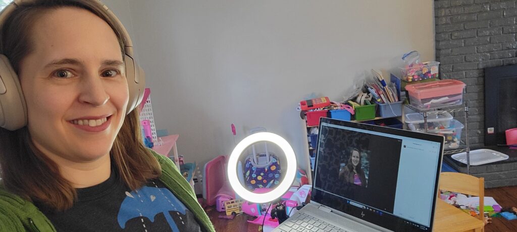 Sara smiles next to her laptop which shows a Zoom meeting. She's wearing headphones and a small circle light shines on her face. In the background are children's toys and art supplies.