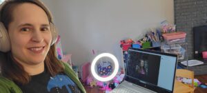Sara smiles next to her laptop which shows a Zoom meeting. She's wearing headphones and a small circle light shines on her face. In the background are children's toys and art supplies.