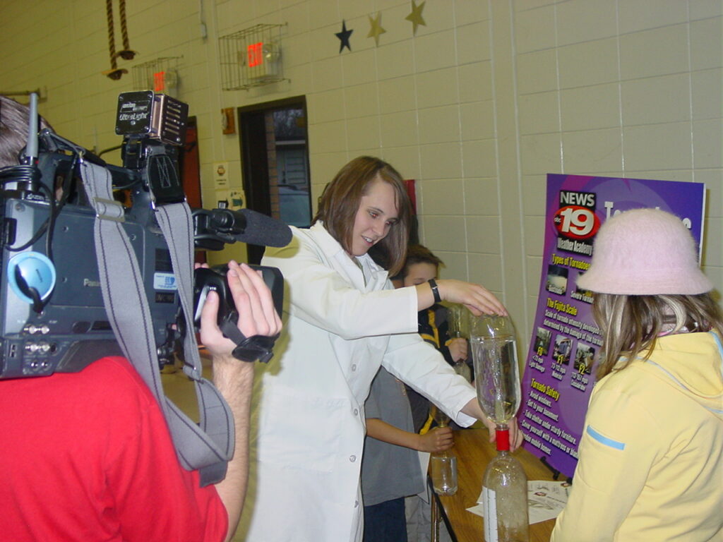 Sara has a camera person filming her as she demonstrates how to make a tornado in a bottle for a group of children. She is wearing a white lab coat and there is a poster with a sign indicating this is an event for WXOW-19 Weather Academy.