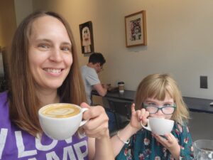 Sara and her daughter hold up mugs with hot drinks (cappuccino with latte art for Sara and a hot chocolate for her daughter whose face is partially obscured as she takes a drink with her pinky up).
