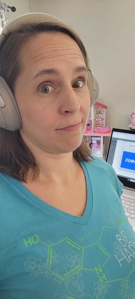 Sara stands in front of her laptop which displays a Zoom meeting sign-in page. She's wearing a blue shirt with the chemical compound for serotonin on it that says, "be happy". She's giving a look to the camera that indicates "this is what you're getting, take it or leave it".