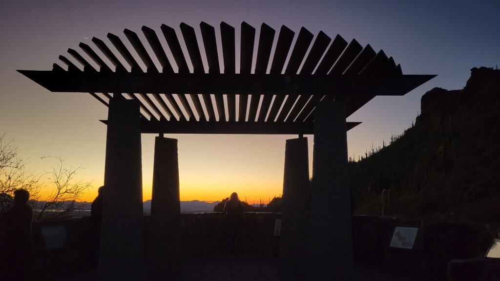 Sunset picture in the mountains of southern Arizona. A sharp hillside on the right shows the outline of saguaro cactus and a pergola dominates the foreground. Mountains are seen in the distance dark purple against the surrounding shades of orange and yellow.
