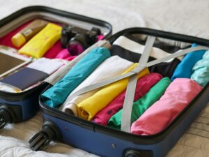 Open blue rolling suitcase filled with neatly rolled clothing on one side and additional supplies including a passport, camera, glasses and lotion on the other side.
