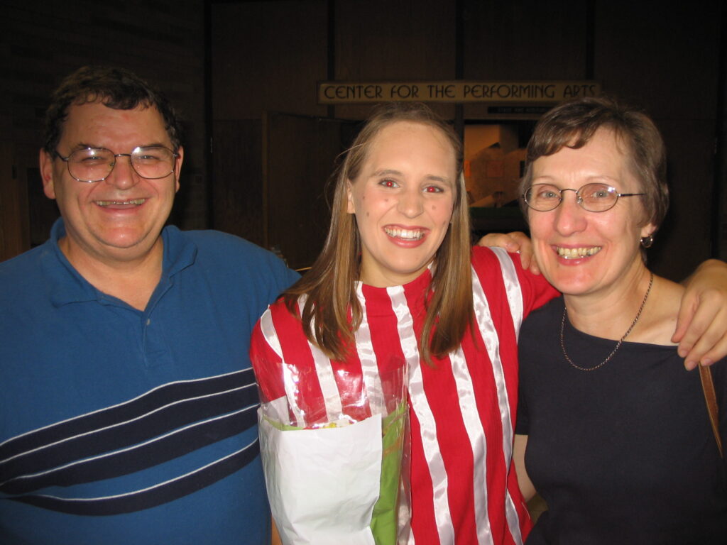 Sara smiles wearing her Peppermint Patty costume and makeup with flowers in her hand.. She stands between her parents, their arms draped around each other.