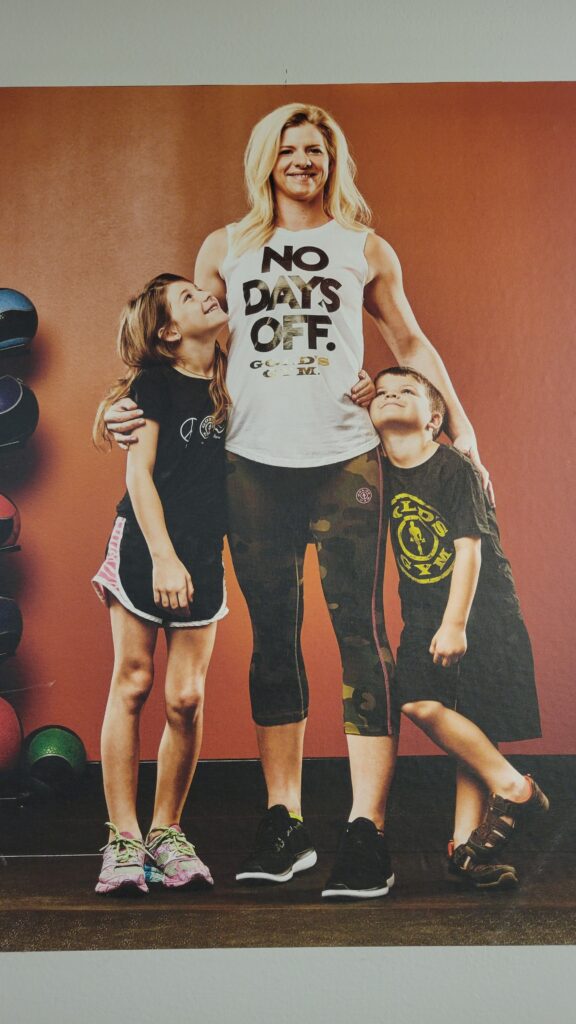 A woman in workout clothes with the phrase "No Days Off" on it stands with her arms draped around a young boy and a young girl who look up at her with admiration. The children, who the image implies are her children, are also wearing workout clothes with the logo of Gold Gym.