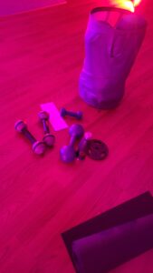 Handheld weights, a water bottle, a yoga mat and a bag of balls sit on a hardwood floor in a room lit in a magenta color.