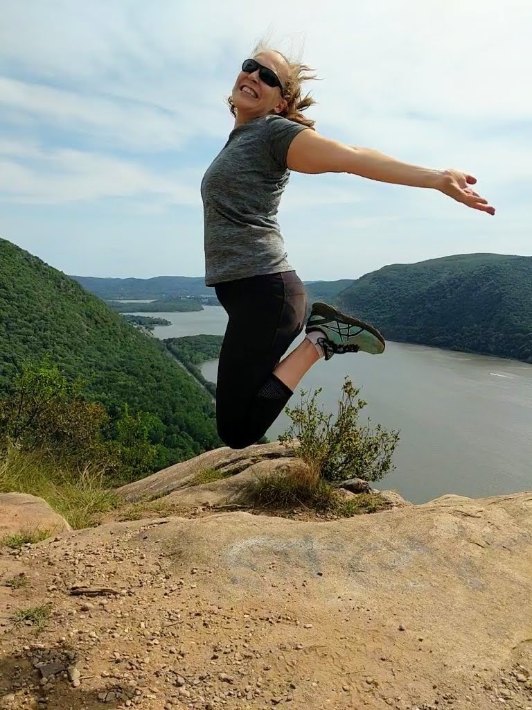 Sara jumping for joy with a scene of a river in a valley behind her. She seems to be floating feet above the ground with her heels tucked in behind her and her arms outstretched.