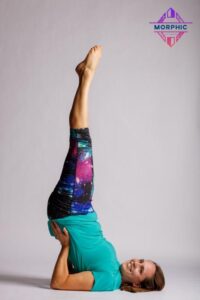Sara wears brightly colored workout clothes. Her face is turned to the camera with a relaxed smile as she is lying balanced on her upper back and neck with her legs extended to the ceiling in the candlestick pose.