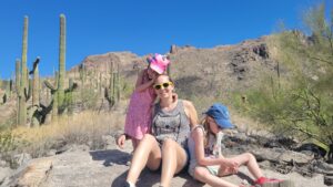 Sara sits on a large flat boulder with her younger daughter hugging her and her older daughter sitting next to her playing with small rocks. The background is desert cactus and shrubs with mountains behind them